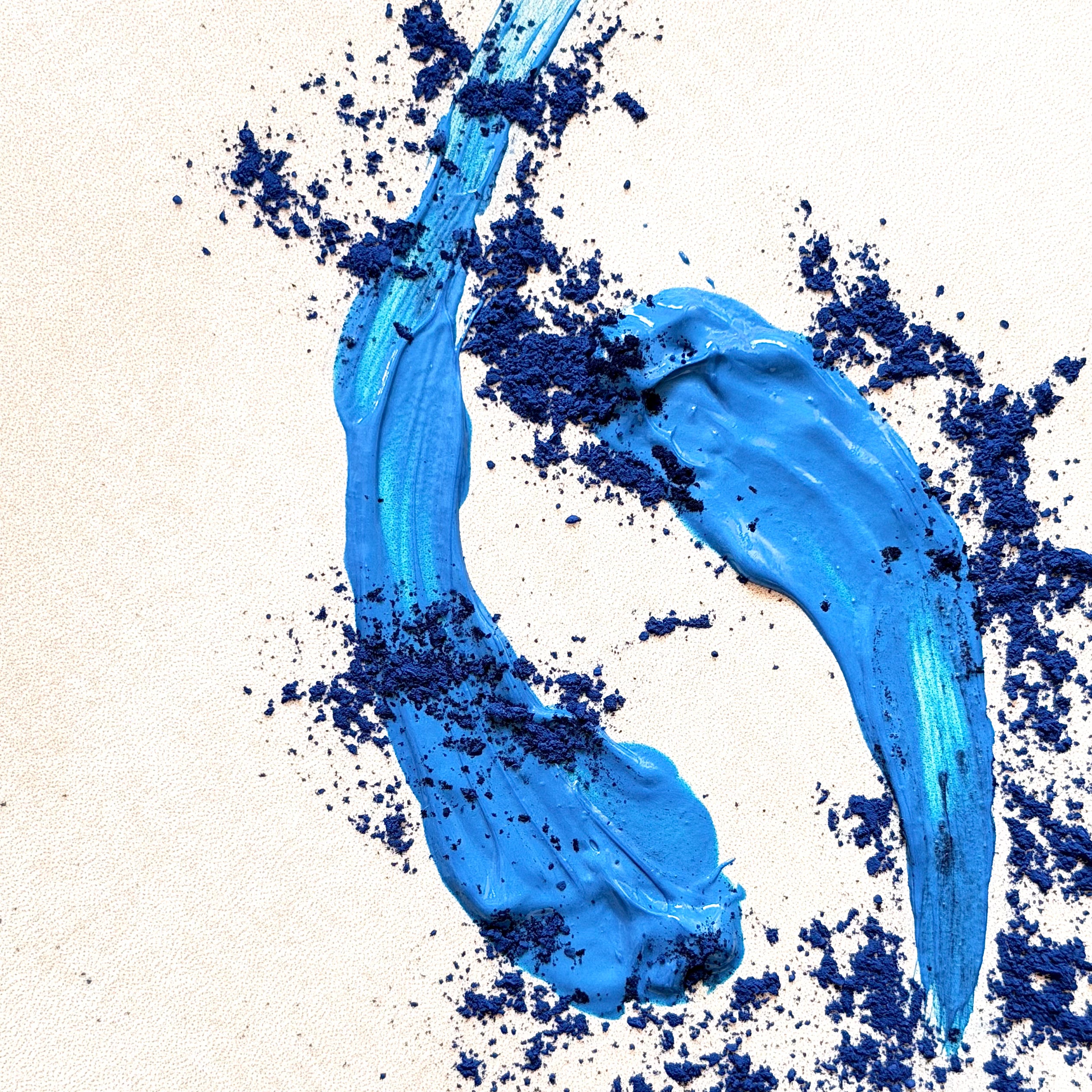 A textured swipe of bright blue cream or gel makeup on a light beige background, surrounded by scattered dark blue powder.